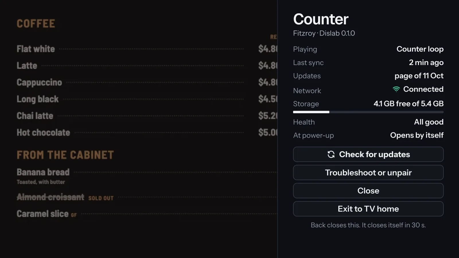 The TV’s menu over a menu board: Playing, Last sync, Updates, Network, Storage, Health and At power-up, then Check for updates, Troubleshoot or unpair, Close and Exit to TV home.